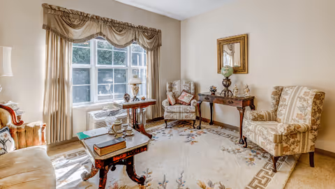 A cozy living room with a large window letting in natural light. The room features two patterned armchairs, a wooden side table with a globe and decorative items, a coffee table with books and a lamp, and a beige sofa. The floor is covered with a light-colored rug with floral and geometric patterns. The walls are painted a light beige, and there is a framed mirror hanging above the side table.