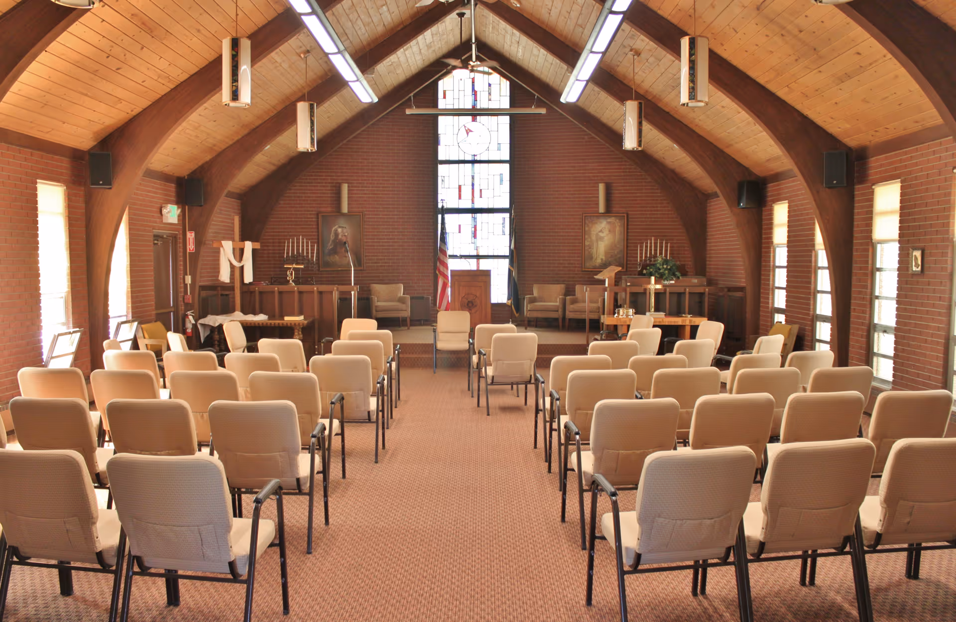 Interior view of a chapel or meeting room with rows of beige chairs facing a wooden podium and stained glass window. The room has a high vaulted wooden ceiling with exposed beams, brick walls, and several framed pictures and candles on the walls.