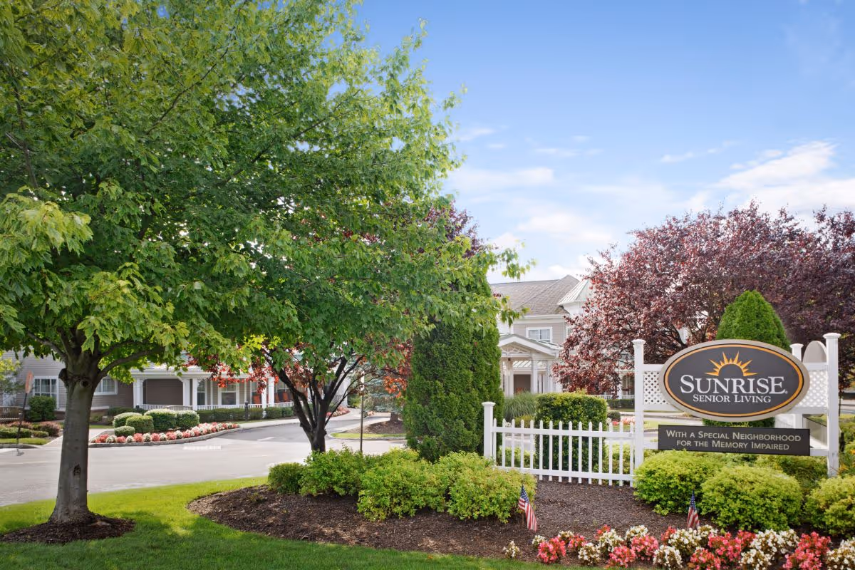 Exterior view of Sunrise Senior Living facility with well-maintained landscaping including green trees, bushes, and colorful flowers. A white fence and a sign reading 'Sunrise Senior Living with a special neighborhood for the memory impaired' are visible in the foreground under a partly cloudy blue sky.