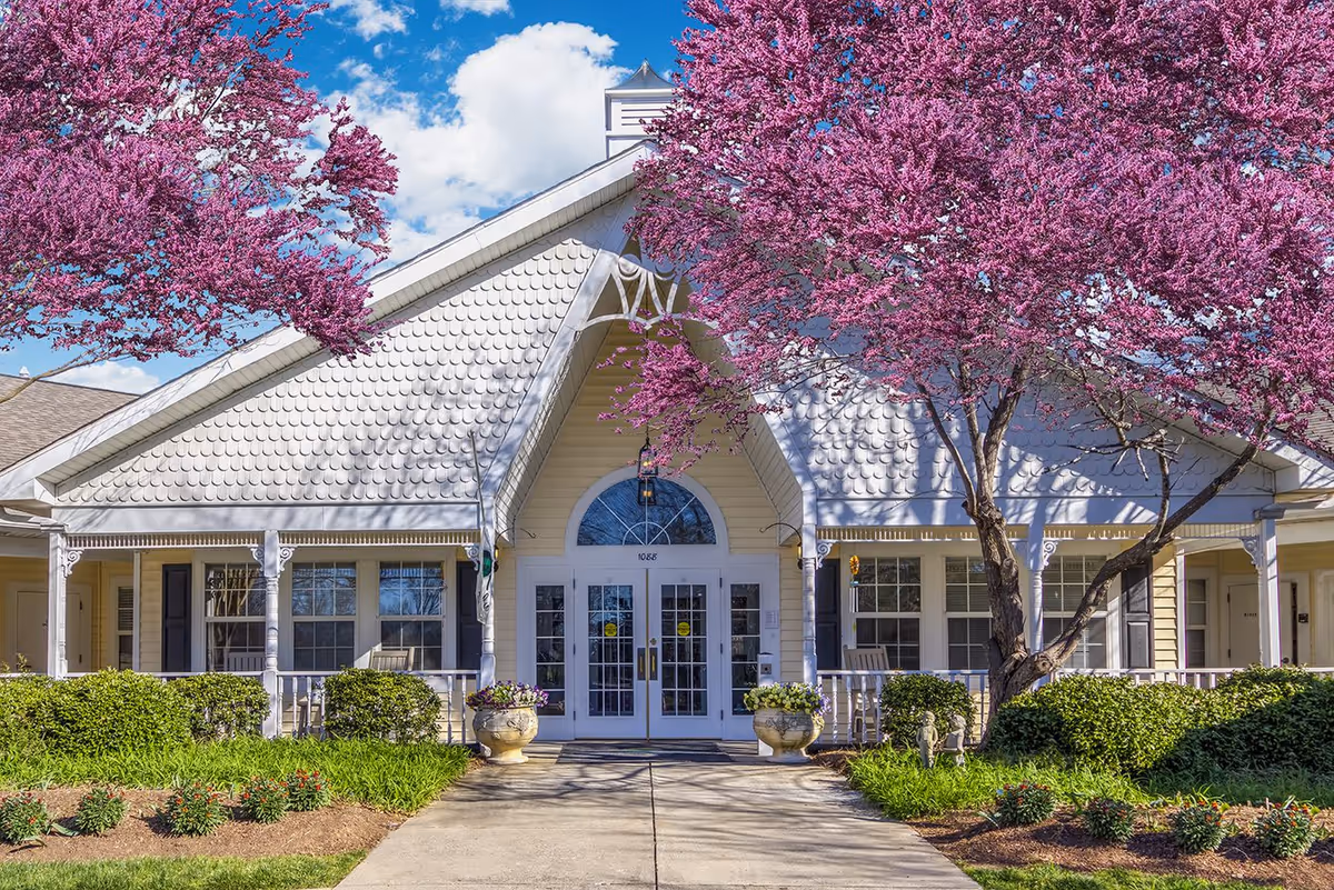 Front exterior view of a senior living facility building with white double doors, a peaked roof, and decorative white siding. The entrance is flanked by two large planters with flowers and surrounded by green bushes and blooming trees with vibrant pink flowers under a blue sky with some clouds.