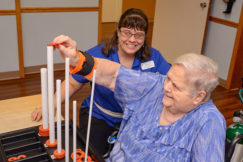 An elderly woman in a wheelchair playing a ring toss game with the assistance of a smiling female caregiver wearing glasses and a blue shirt with a name tag that reads Molly. They are indoors at a wooden table with a light-colored floor and walls in the background.