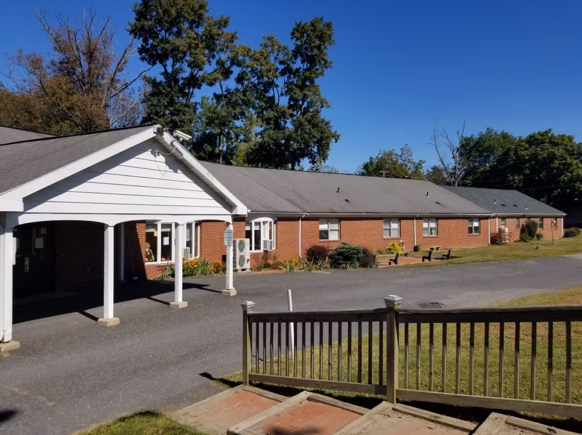 Exterior view of a single-story brick building with a covered entrance and a paved driveway. There are trees and a clear blue sky in the background, and a wooden railing and steps in the foreground.