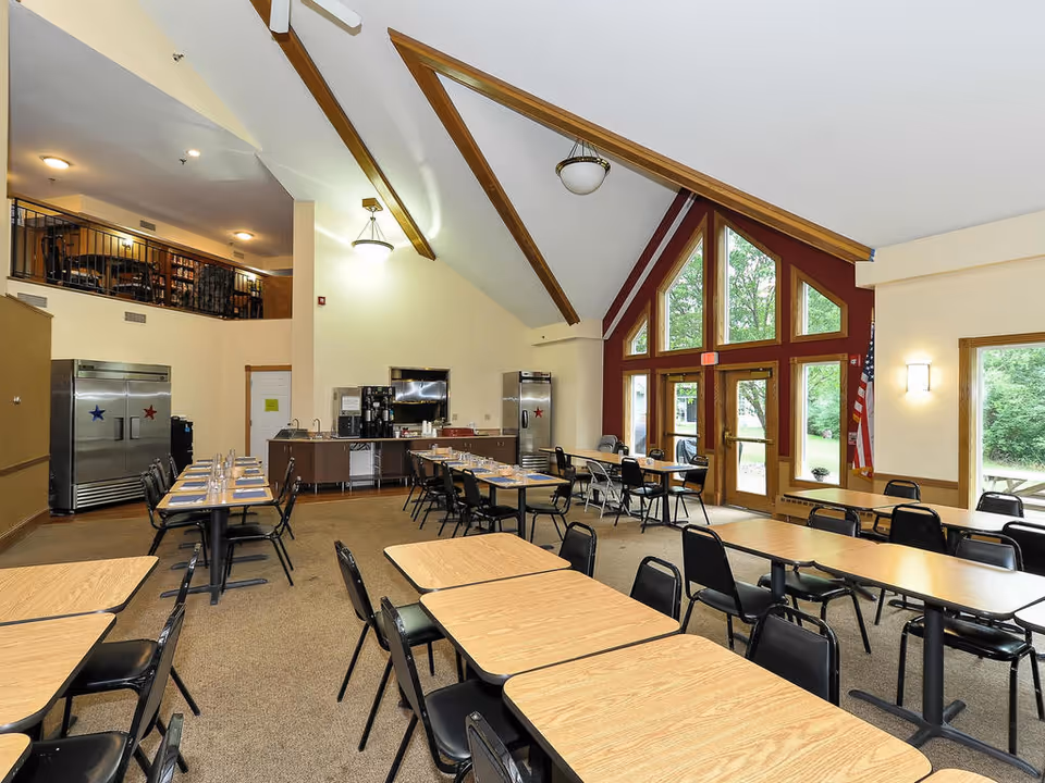 A spacious dining room with multiple rectangular wooden tables and black chairs arranged neatly. The room features a high vaulted ceiling with wooden beams and large triangular windows letting in natural light. There are two large stainless steel refrigerators with star decals, a coffee station, and an American flag near the glass doors that lead outside to a green area with picnic tables.