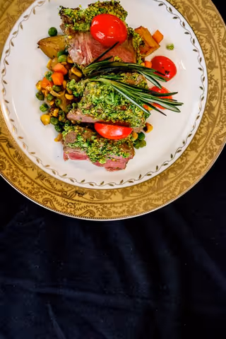 A gourmet plate featuring slices of herb-crusted meat garnished with cherry tomatoes and fresh rosemary, served with a side of mixed vegetables including peas, carrots, and corn on an ornate white and gold-rimmed plate.