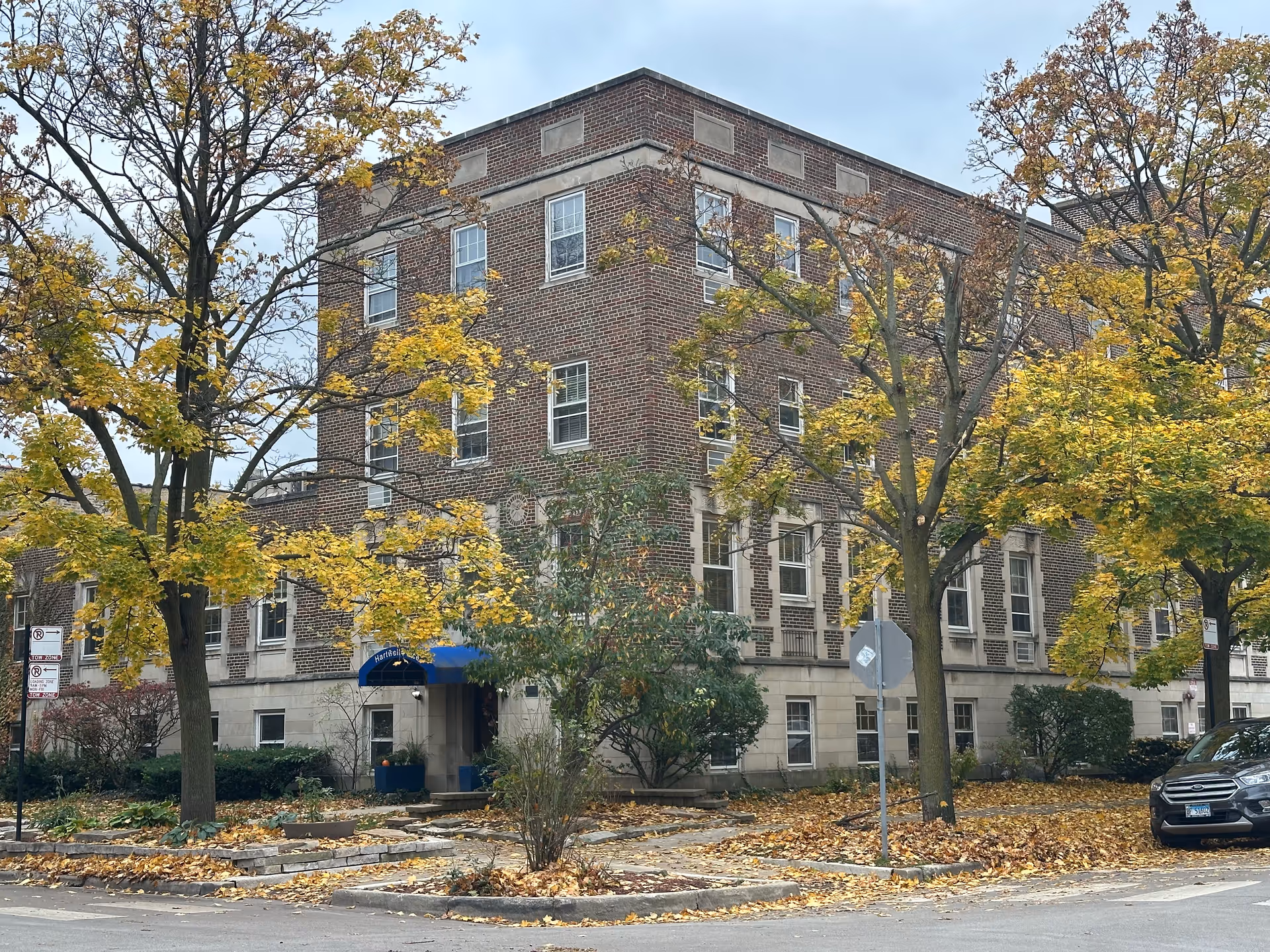 Exterior view of a multi-story brick building with several windows, surrounded by trees with yellow autumn leaves. There is a blue awning over the entrance that reads 'Hartwell Place'. A car is parked on the street nearby, and fallen leaves cover the ground.