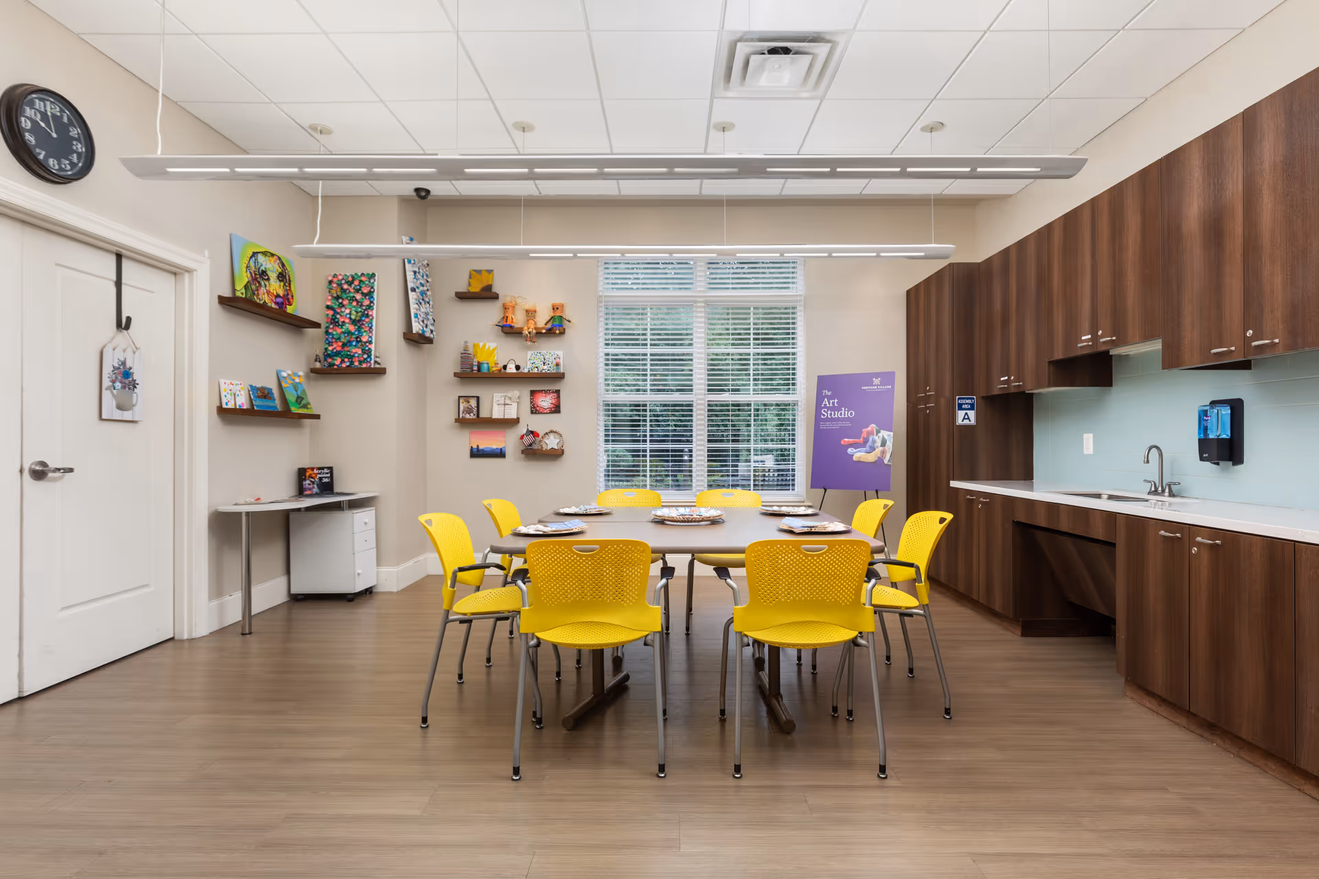 A bright art studio room with a large rectangular table surrounded by eight yellow chairs. The room has wooden cabinets and a sink on the right side, shelves with colorful artwork on the left wall, and a large window with blinds in the center background. A clock is mounted on the left wall above a white door, and a purple sign reading 'The Art Studio' is visible near the window.