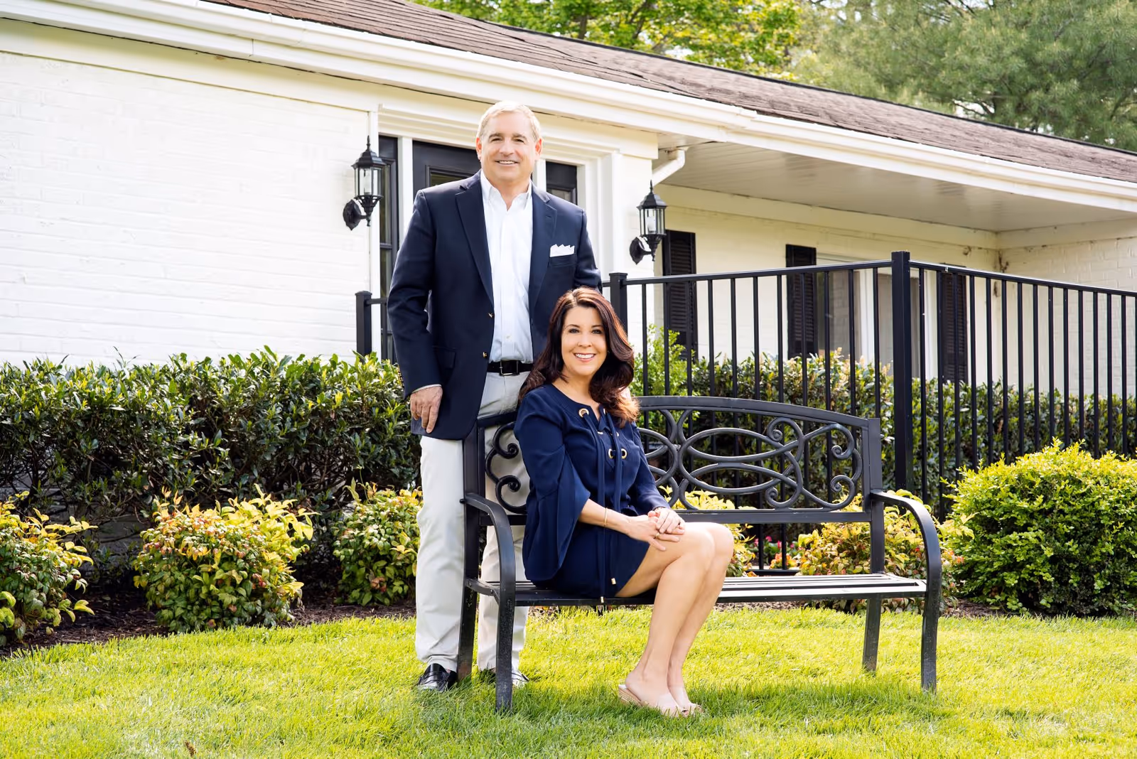 A man in a dark blazer and light pants stands behind a woman in a navy dress sitting on a black metal bench on a grassy lawn. Behind them is a white brick building with black shutters and a black metal railing, surrounded by green bushes and trees.