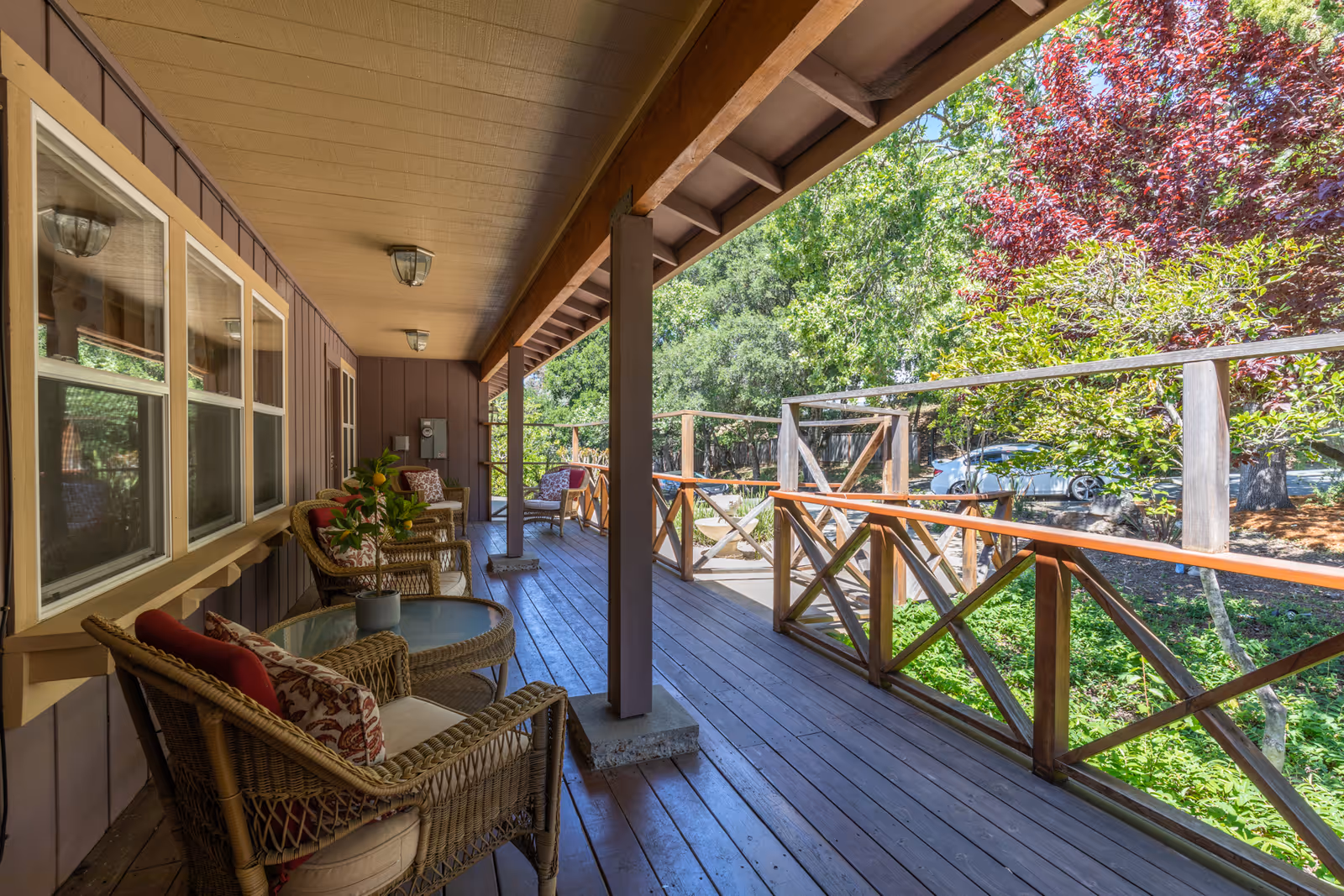 A covered wooden porch with wicker chairs and a table, wooden railing, and trees visible beyond.