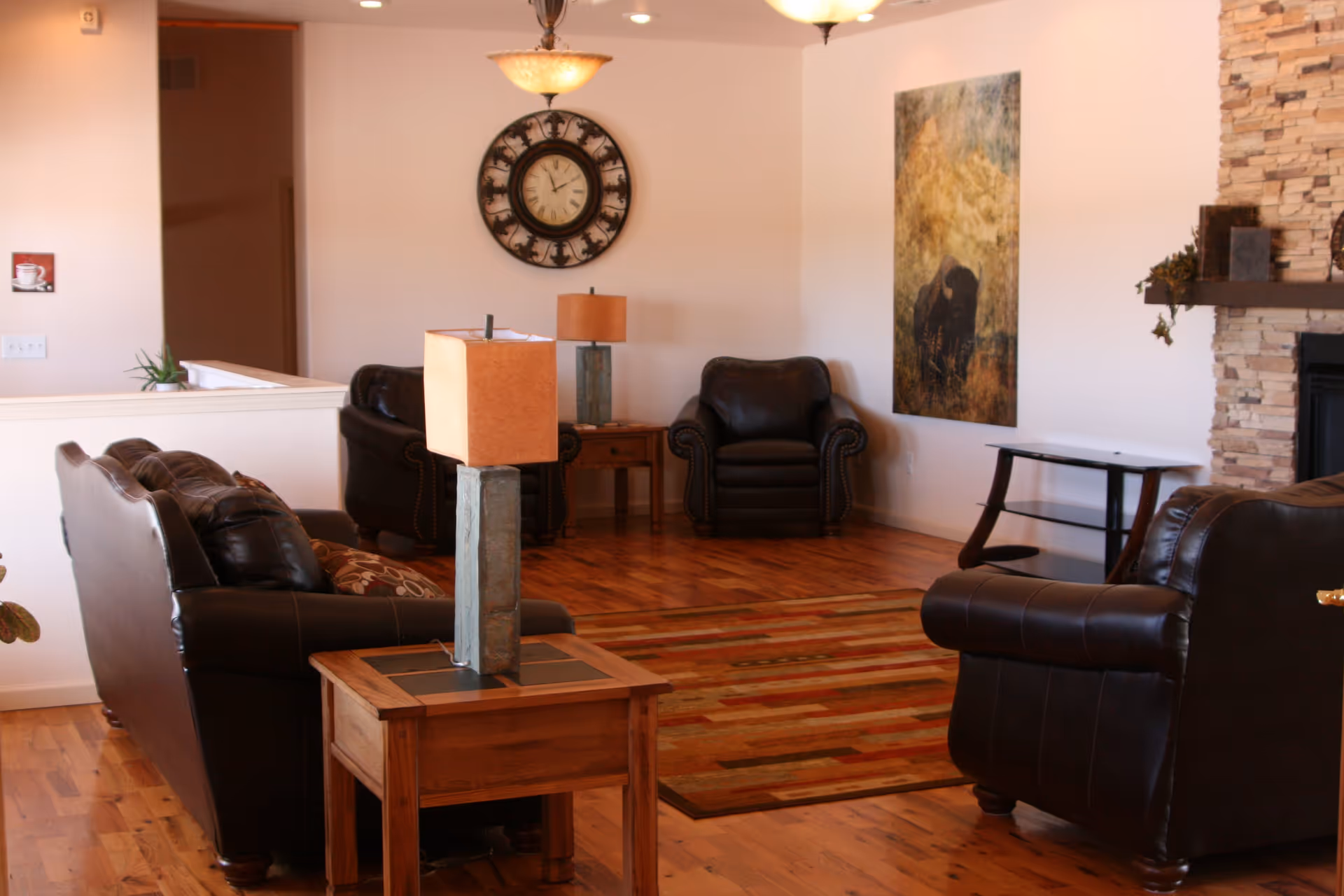 A cozy living room with leather sofas and armchairs, wooden side tables and lamps, a rug, wall clock, artwork, and a stone fireplace.
