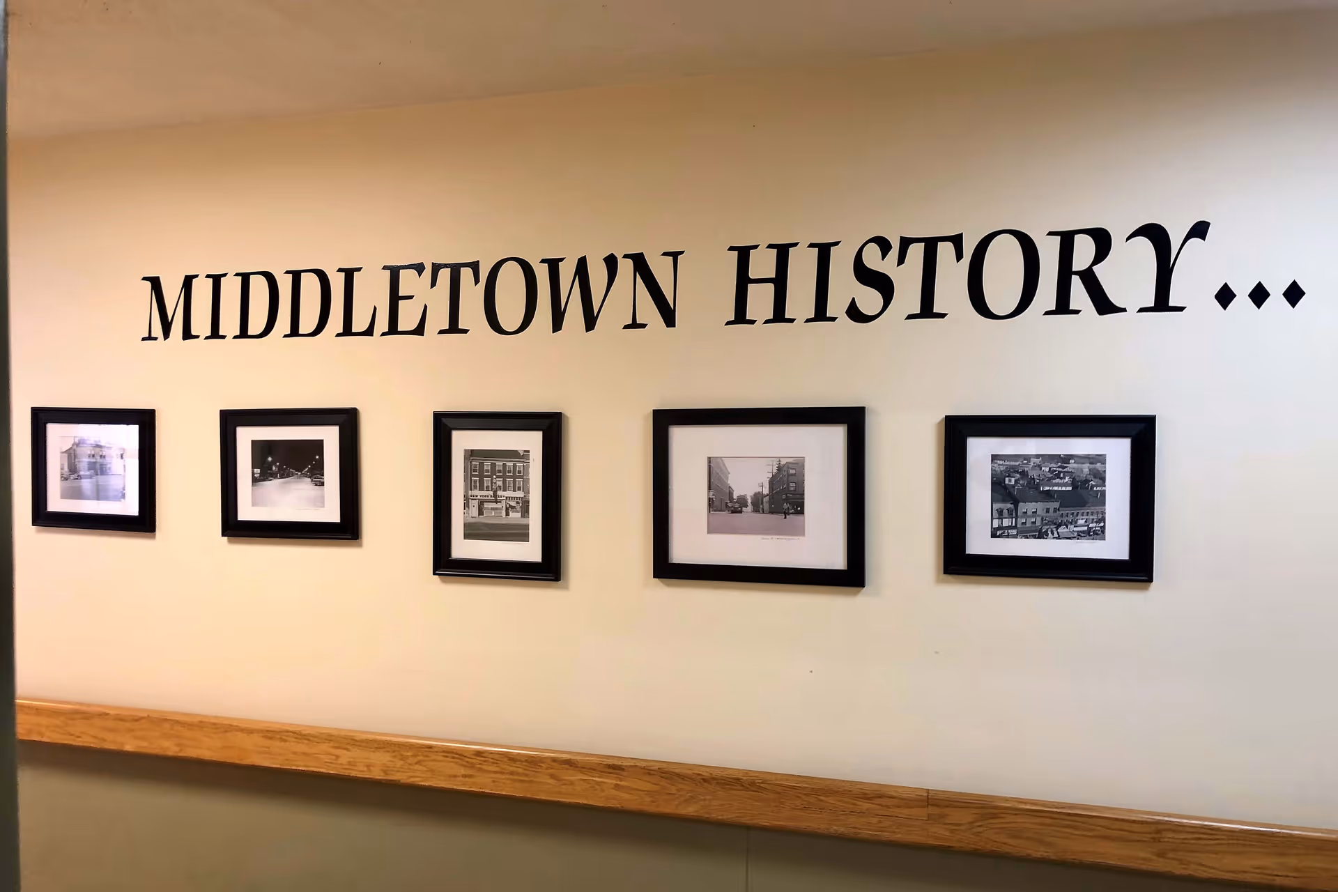 A beige wall with the words 'MIDDLETOWN HISTORY...' written in large black letters. Below the text, there are five framed black and white historical photographs arranged in a row. A wooden handrail runs along the wall beneath the pictures.