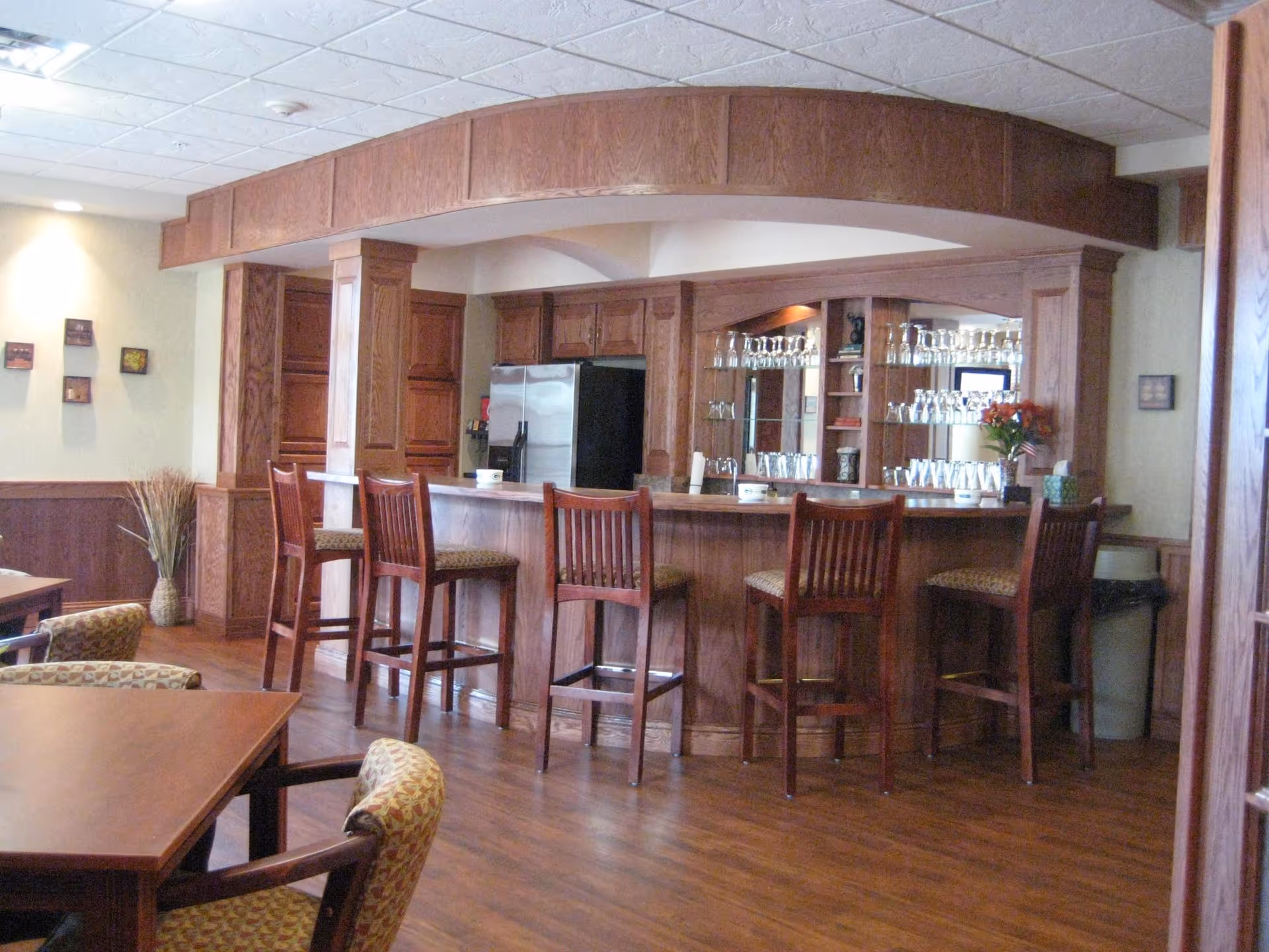 Interior view of a retirement community dining area featuring a wooden bar with five high chairs, a mirrored back wall with shelves holding glassware, and a stainless steel refrigerator. The room has wooden flooring, beige walls, and several tables and chairs in the foreground.