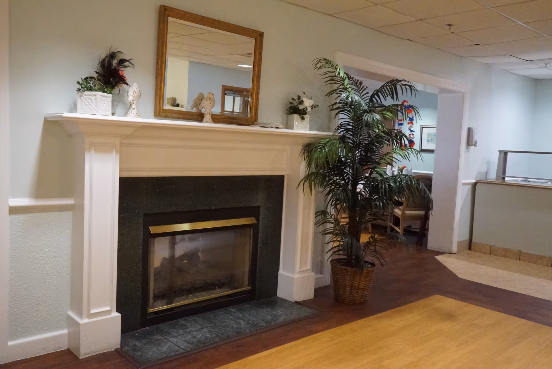 Interior view of a senior living facility featuring a white fireplace with a black hearth and a gold-framed mirror above it. Decorative items including plants and figurines are placed on the mantel. To the right, there is a large potted plant and a glimpse into a dining area with tables and chairs. The floor has a combination of wood and tile, and the walls are painted light blue.
