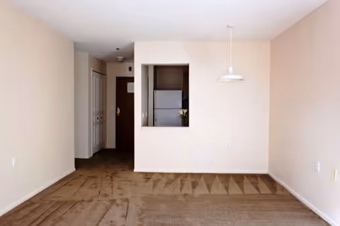 Empty room with beige walls and brown carpet, featuring a small pass-through window to a kitchen area with a white refrigerator. A white hanging light fixture is above the carpeted floor area, and a hallway with closed doors is visible in the background.