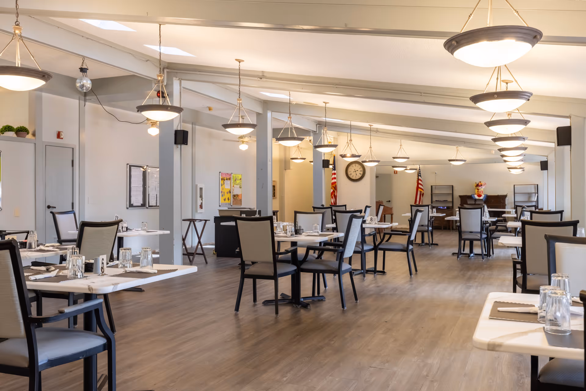 A spacious dining room in a senior living facility with multiple tables and chairs arranged neatly. Each table is set with placemats, napkins, and upside-down drinking glasses. The room features wooden flooring, hanging ceiling lights, and two American flags near the back wall. A large clock is mounted on the wall, and there are bulletin boards and shelves in the background.