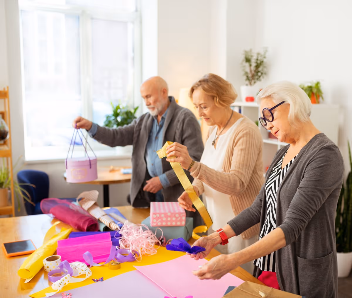 Three elderly individuals standing around a table covered with colorful wrapping paper, ribbons, and gift boxes, engaged in gift wrapping in a bright room with large windows and plants.