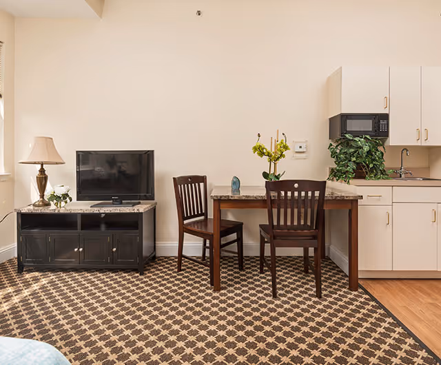 Interior view of a senior living facility room featuring a small dining table with two wooden chairs, a black TV stand with a flat-screen television, a table lamp, and a kitchenette area with white cabinets, a microwave, and a sink. The floor is covered with a patterned carpet and part of the floor near the kitchenette is wooden.