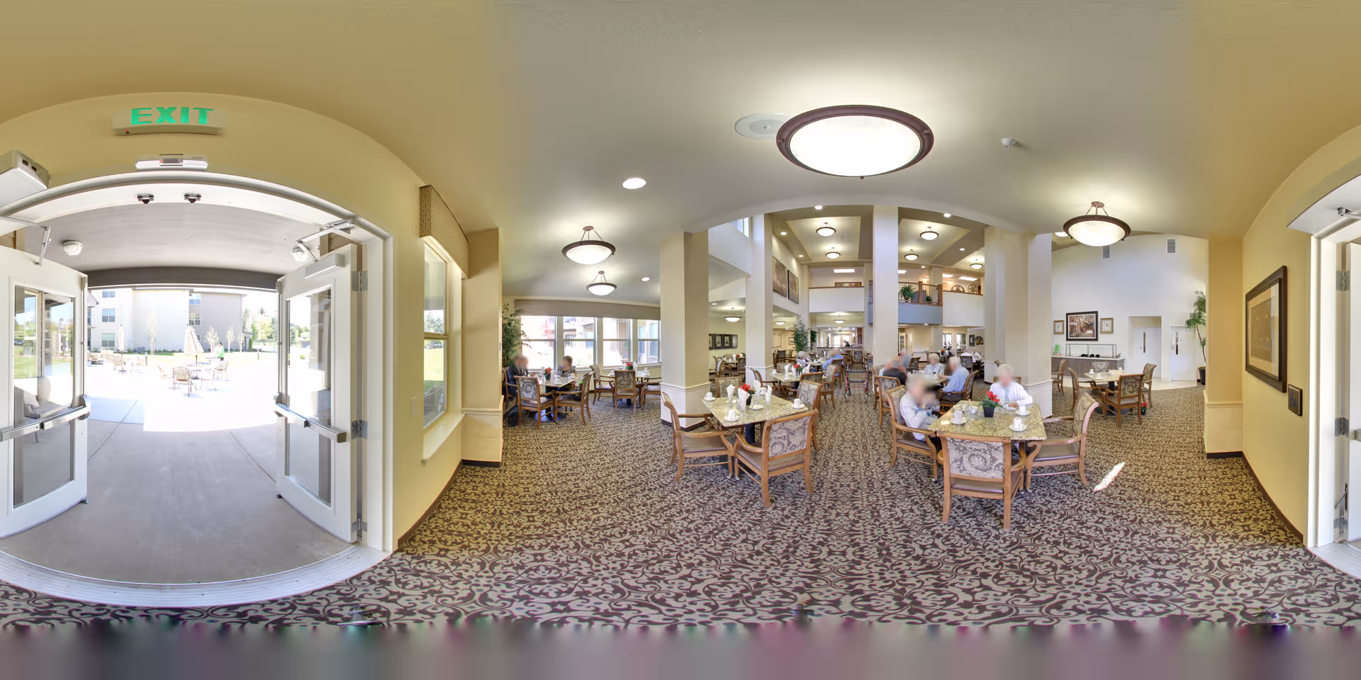 Interior view of a spacious dining area in a senior living facility with multiple tables and chairs. Several elderly residents are seated at the tables, engaging in conversation and dining. The room features large windows allowing natural light, patterned carpet, and ceiling lights. Double doors lead outside to a patio area with outdoor seating and umbrellas.