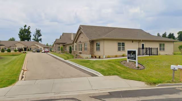 Exterior view of a single-story senior living facility named Willowick Moments, showing a paved driveway, green lawns, and multiple connected beige buildings with brown roofs under a cloudy sky.