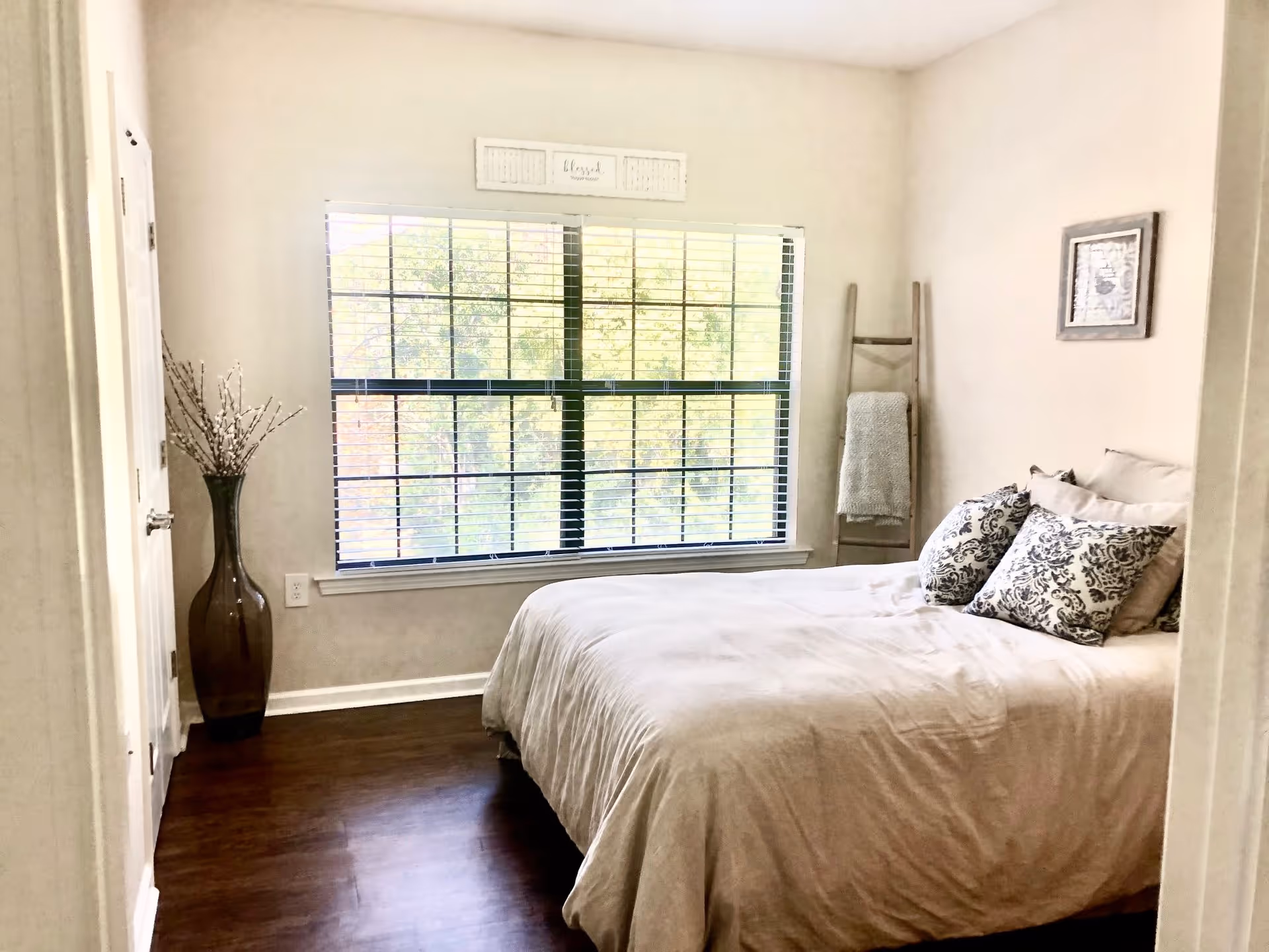 A bright bedroom with a large window covered by horizontal blinds. The room features a bed with beige bedding and decorative pillows with a black and white pattern. A wooden ladder with a gray blanket draped over it leans against the wall, and a tall brown vase with decorative branches is placed near the door. The walls are light-colored, and there is a framed picture hanging on the wall above the bed.