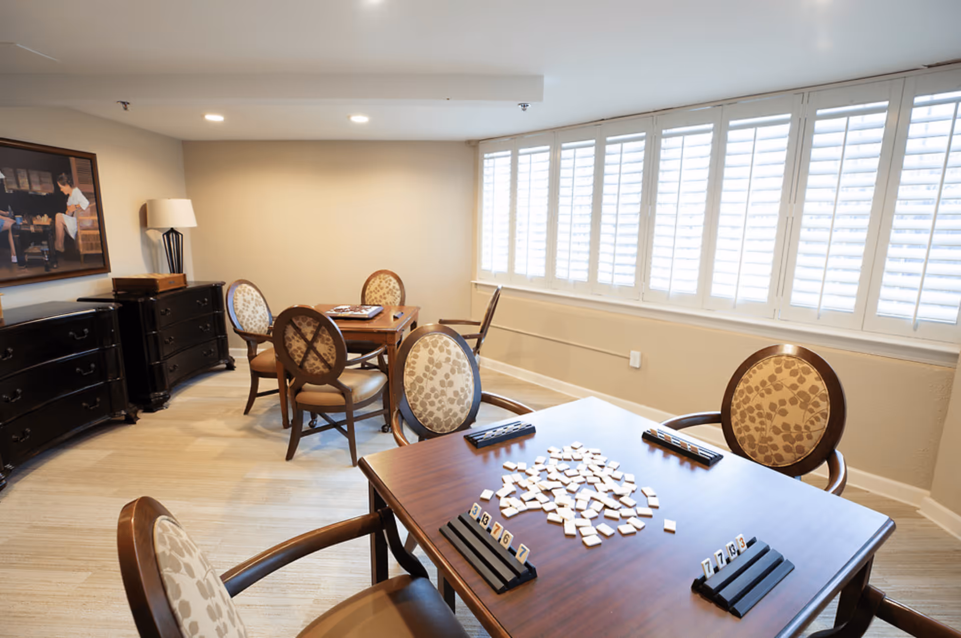 A bright room with two wooden tables and six upholstered chairs arranged around them. One table has a game set up with tiles and racks, likely for a tile-based game. The room has large windows with white shutters, light-colored walls, and two black dressers with a lamp and framed artwork on the wall.