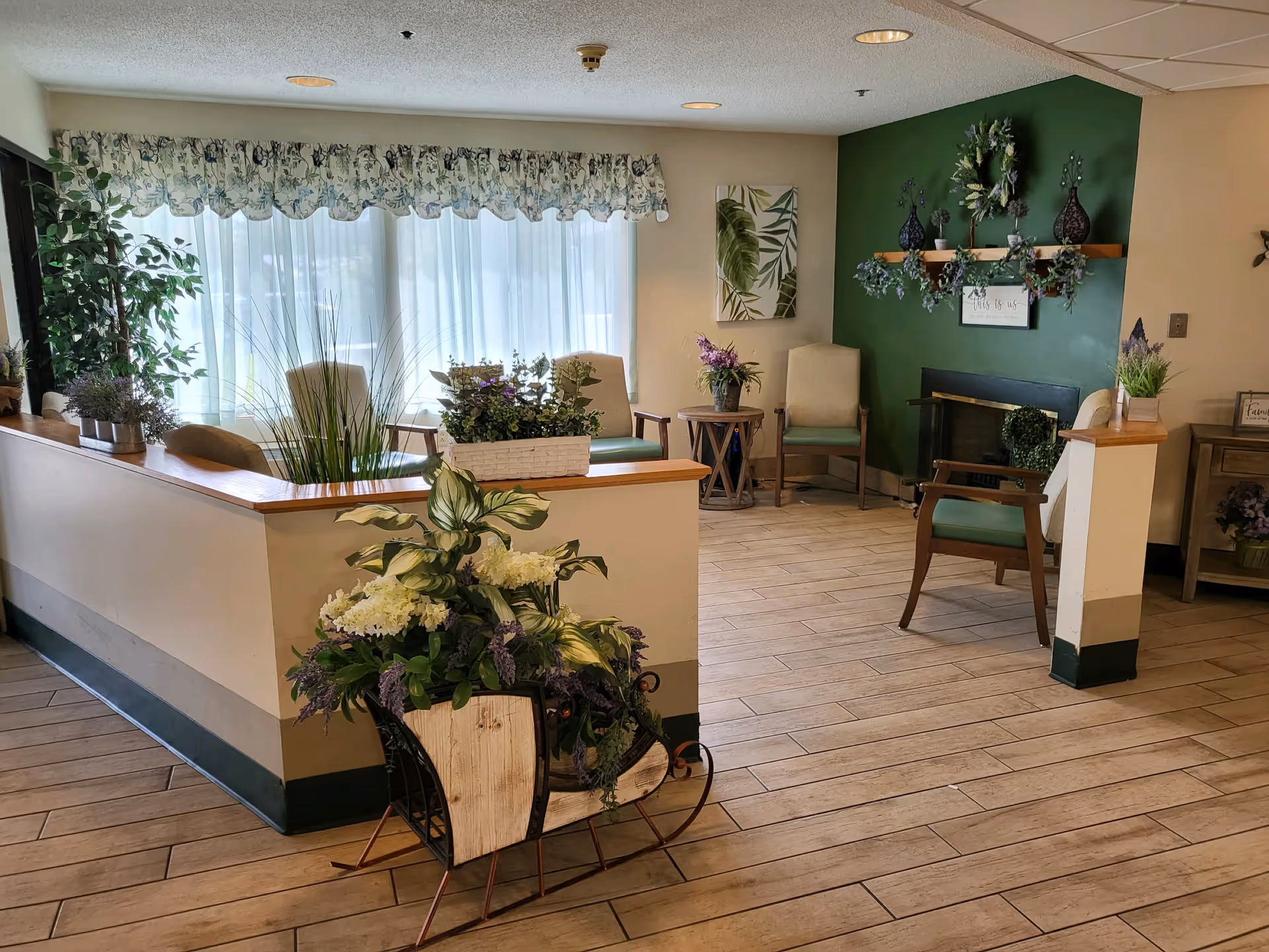 A cozy sitting area in a senior living facility with wooden flooring, several chairs with green cushions, a green accent wall with decorative shelves and plants, a fireplace, and a large window with floral curtains allowing natural light to fill the room. There are various potted plants and floral arrangements placed around the space, creating a welcoming atmosphere.