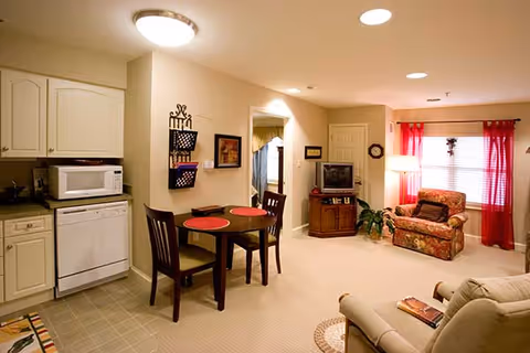 A cozy living area in a nursing center featuring a small dining table with two chairs and red placemats, a kitchen area with white cabinets, a microwave, and a dishwasher, a comfortable armchair with floral upholstery near a window with red curtains, a TV on a wooden stand, and a beige recliner chair in the foreground.