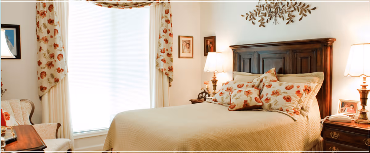A cozy bedroom with a large wooden headboard bed adorned with floral patterned pillows and a beige quilt. The room features floral curtains framing a large window with blinds, two bedside tables with lamps, framed pictures on the walls, and a comfortable armchair next to a wooden desk.