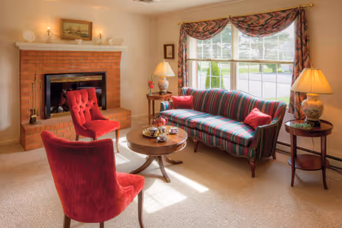 Cozy living room with a striped sofa, two red upholstered chairs, a round wooden coffee table, a brick fireplace, and a window with patterned curtains.