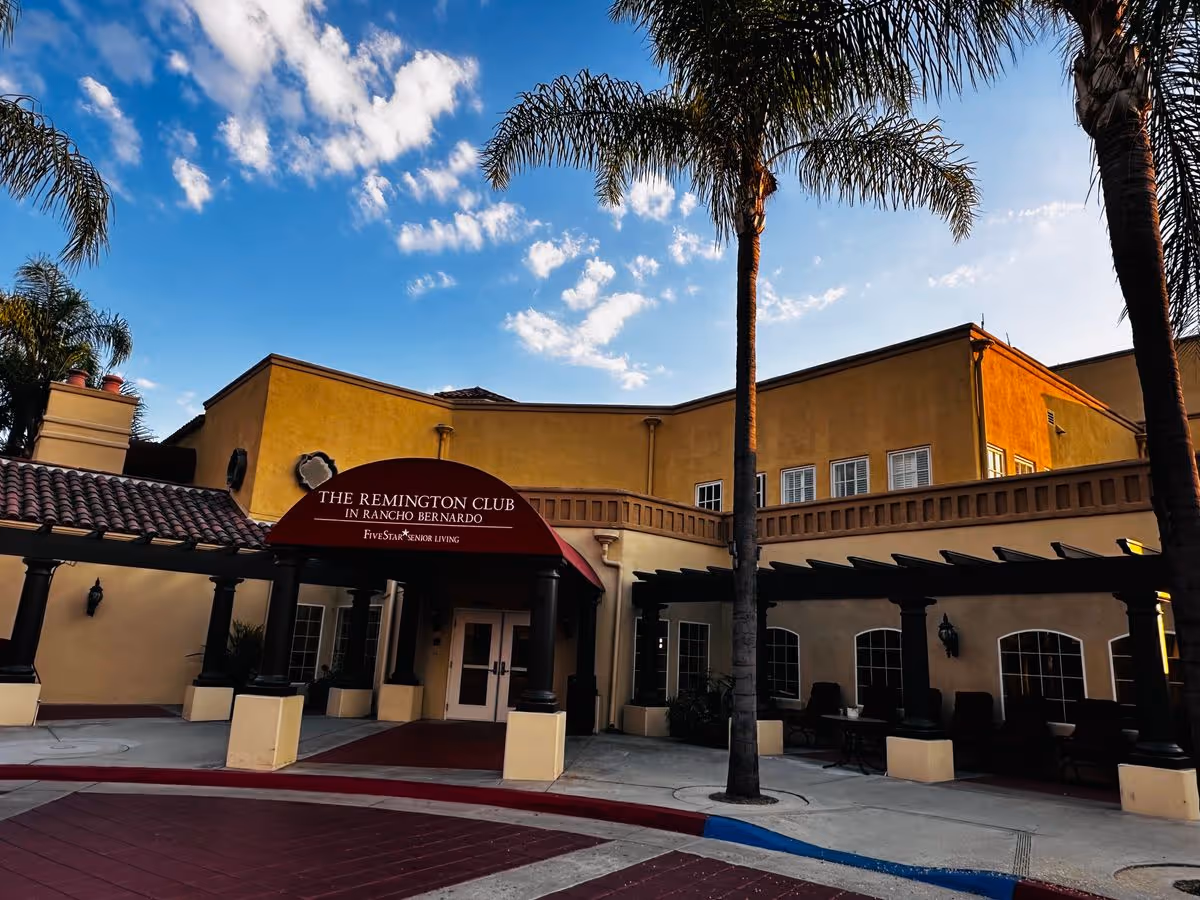 Entrance of The Remington Club senior living building with a red awning, palm trees, and yellow stucco under a blue sky.