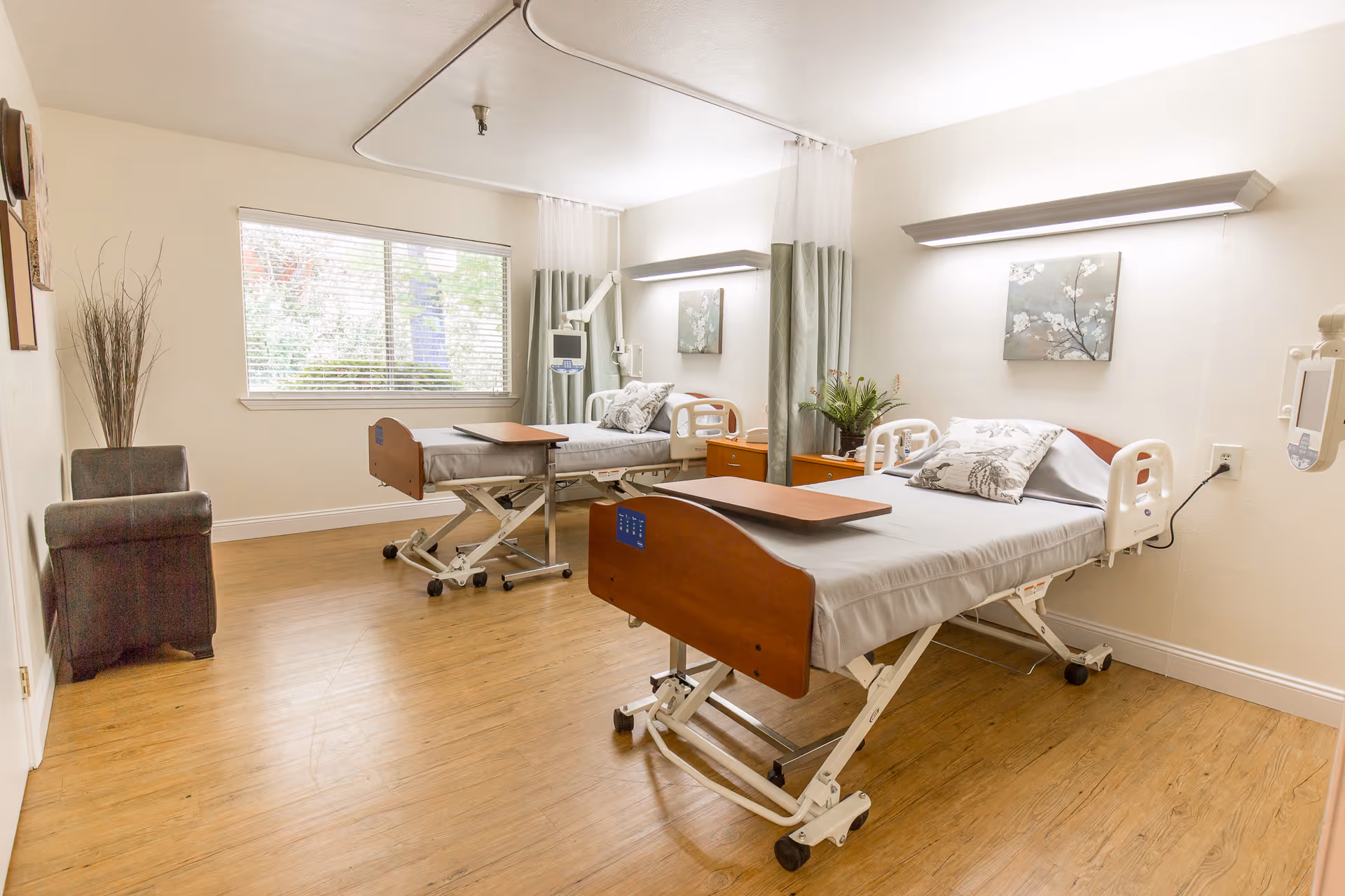 A bright and clean room with two hospital-style beds, each equipped with a pillow and a small overbed table. The room has wooden flooring, a large window with blinds, a small armchair, and decorative plants and artwork on the walls. Privacy curtains are partially drawn between the beds.