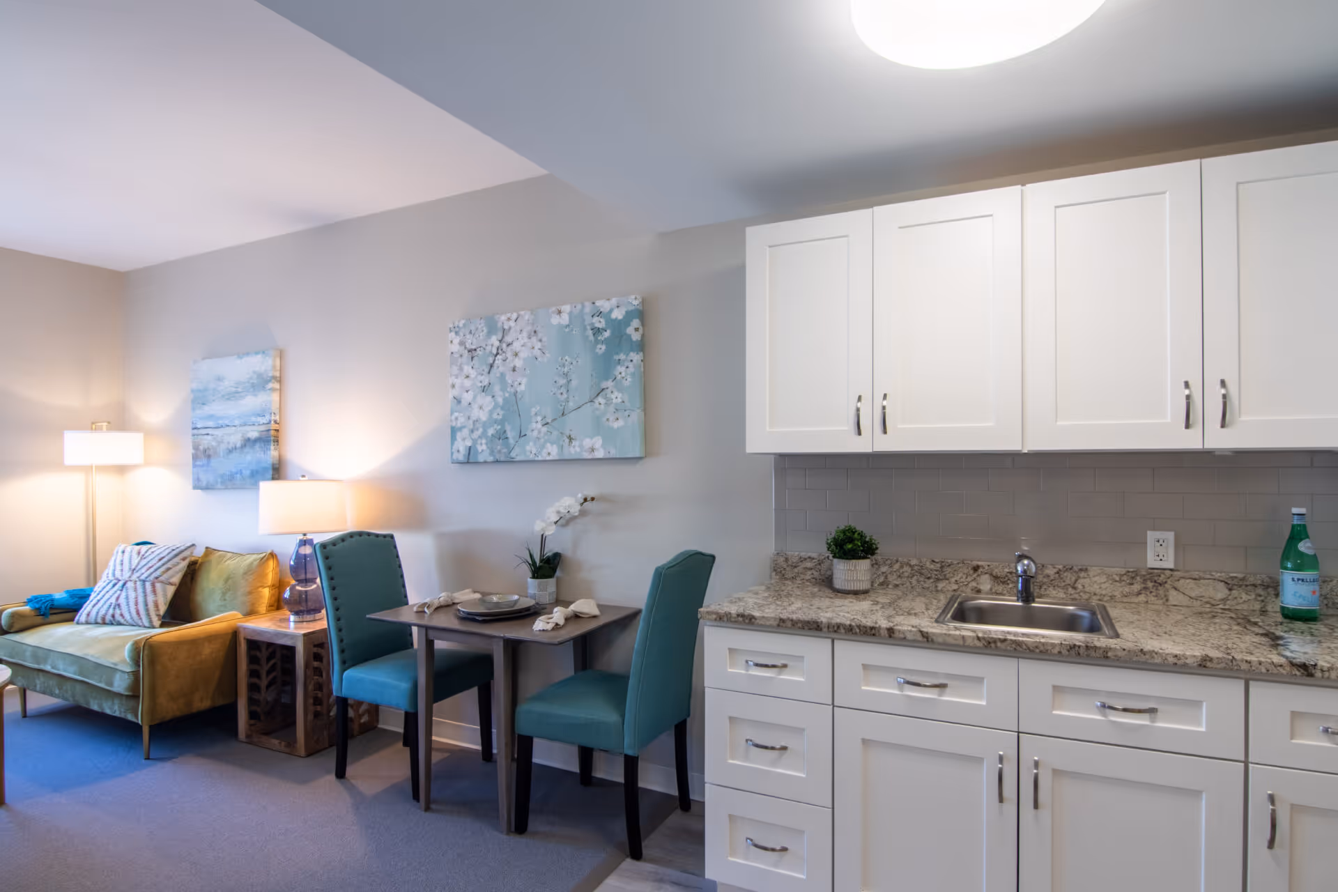 A cozy living area and kitchenette in a senior living facility. The living area features a green velvet sofa with decorative pillows, a wooden side table with a lamp, and two teal upholstered chairs around a small dining table set with plates and napkins. The kitchenette has white cabinets, a granite countertop, a stainless steel sink, a small potted plant, and a bottle of sparkling water. The walls are decorated with two paintings, one depicting a floral design and the other a landscape.