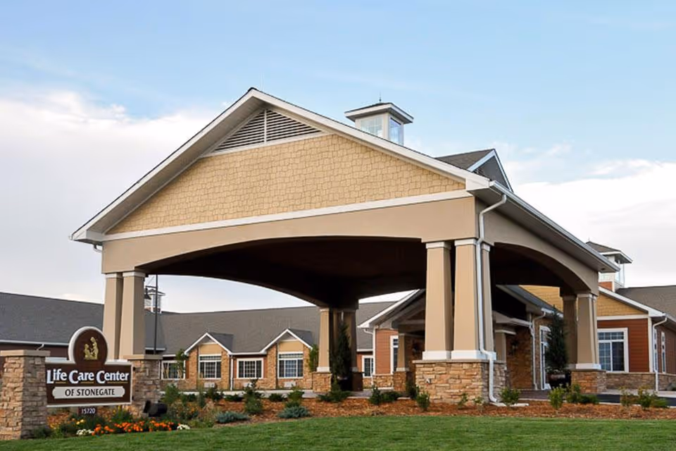 Exterior view of Life Care Center of Stonegate showing a large covered entrance with stone pillars, landscaped greenery, and a clear sky in the background.