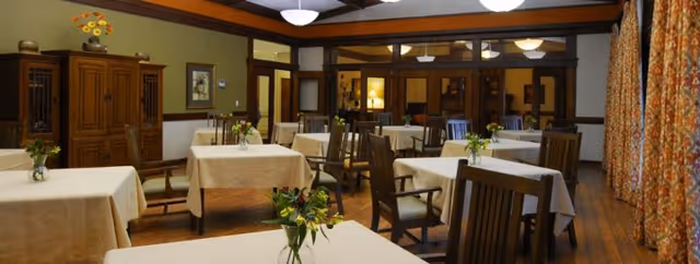 A dining room with multiple tables covered in white tablecloths, each adorned with small flower arrangements. Wooden chairs surround the tables, and the room features wooden flooring and wood trim along the walls. There is a wooden cabinet against one wall and patterned curtains on the windows.
