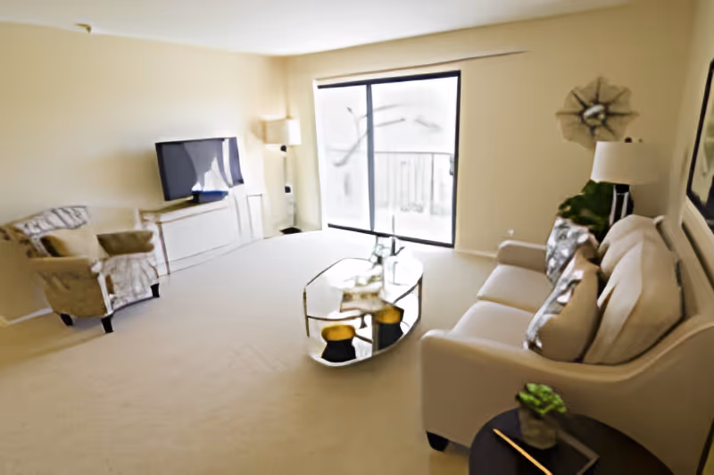 Bright living room with a sofa and pillows, an armchair, wall-mounted TV, glass coffee table, and sliding glass door to a balcony.