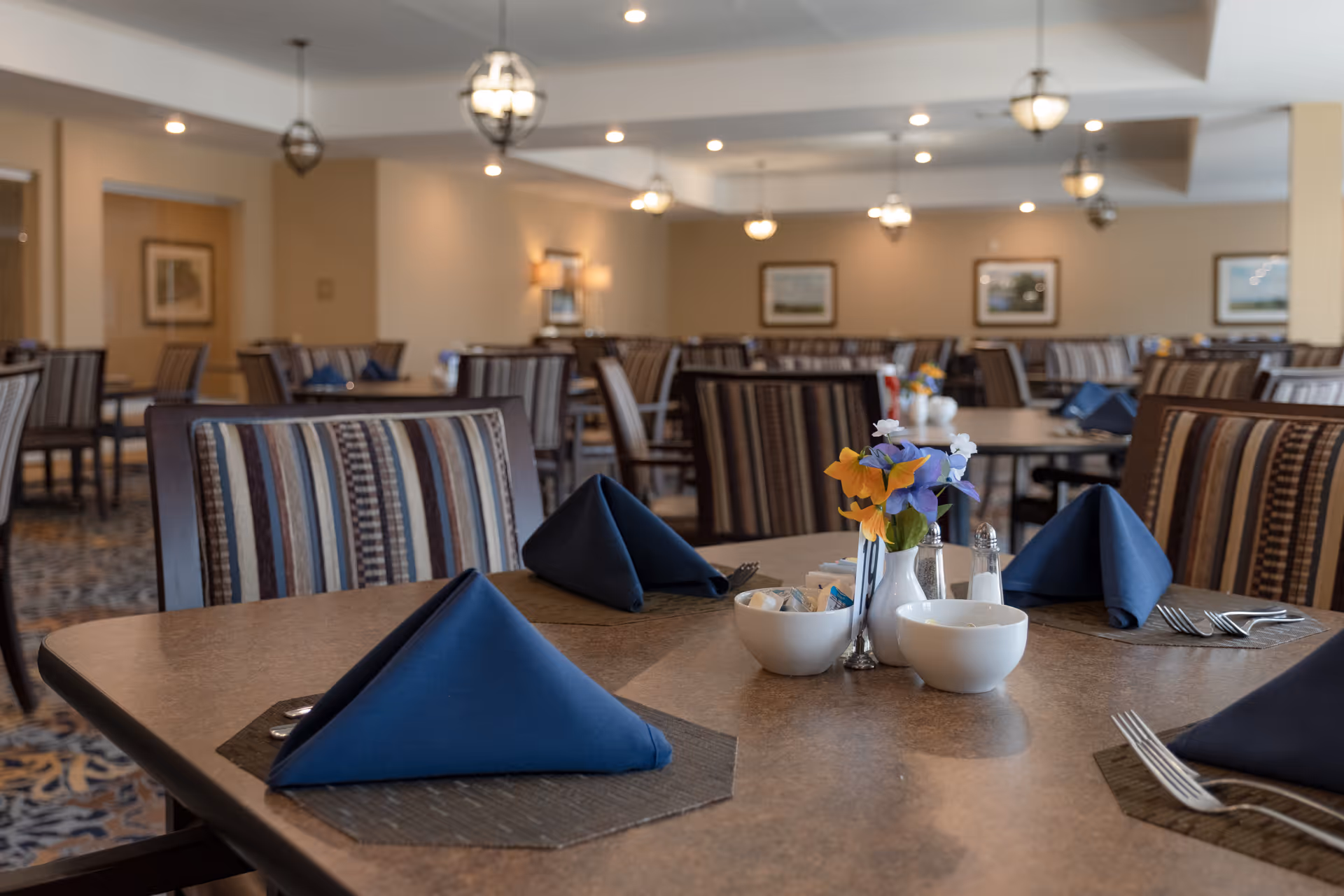 A dining room with multiple tables and chairs. Each table is set with blue folded napkins, silverware, small bowls, and a small vase with colorful flowers. The room has soft lighting with hanging light fixtures and framed pictures on the walls.