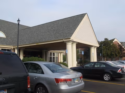 Covered entrance with large columns and several parked cars in the lot in front of a residential building.