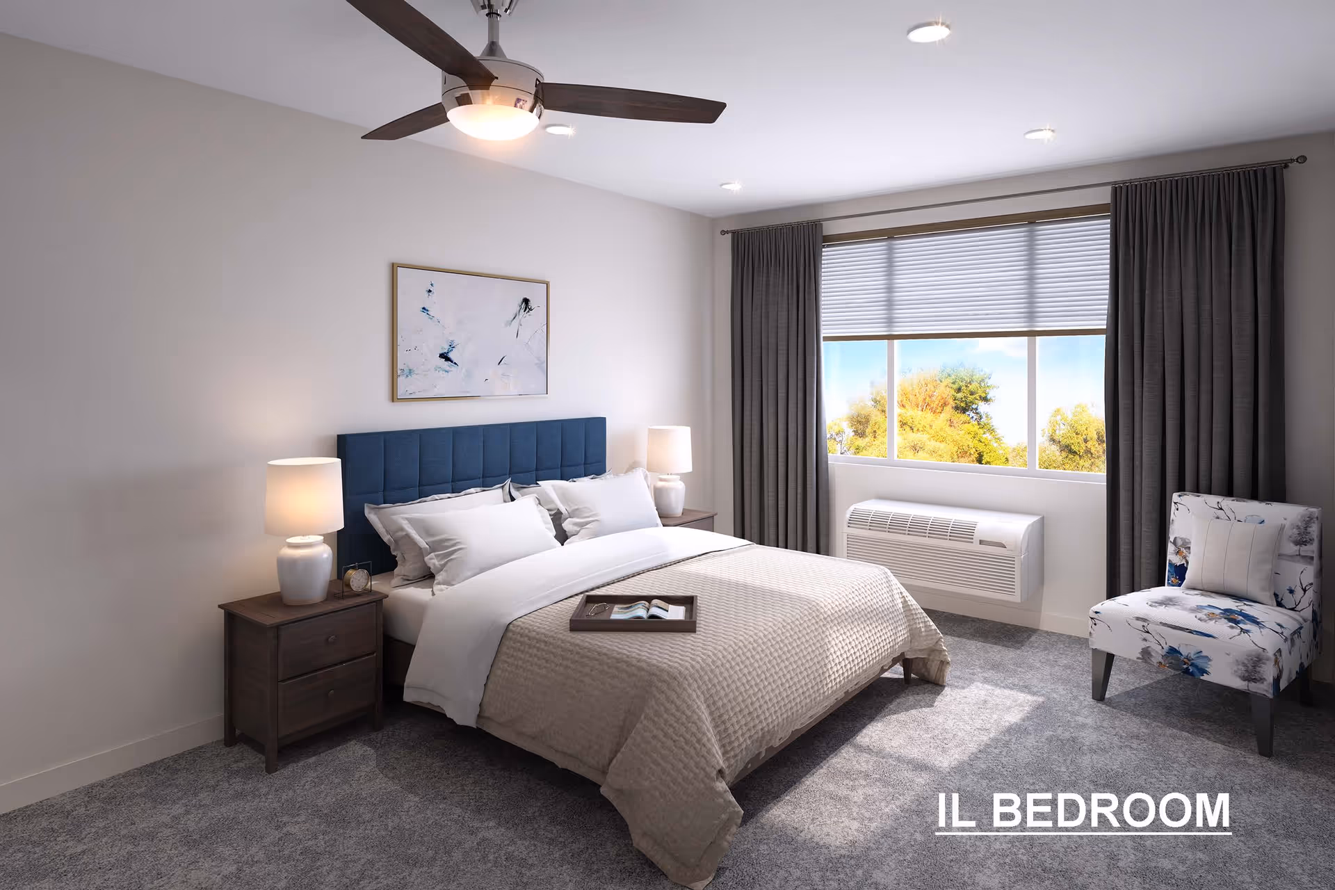 A bright and modern bedroom with a large bed featuring a blue upholstered headboard, white pillows, and a beige quilted blanket. There are two wooden nightstands with white lamps on either side of the bed. A tray with a book and glasses rests on the bed. The room has gray carpet, a ceiling fan with a light, a large window with gray curtains and blinds, and a floral-patterned chair near the window.