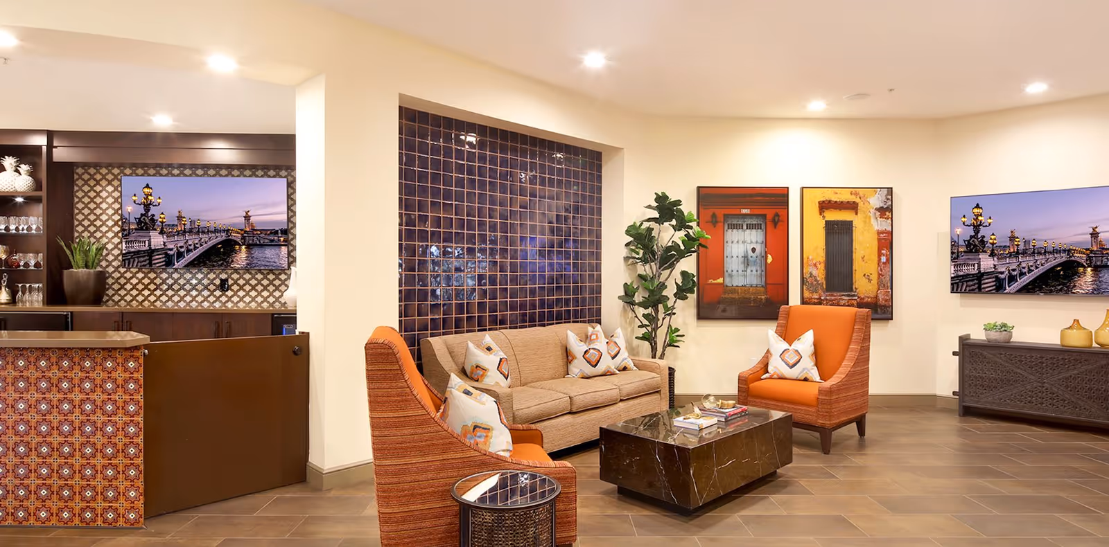 A cozy living room area with a beige sofa and two orange armchairs, each with decorative pillows. The room features a dark marble coffee table, a side table, and a tall green plant. The walls are adorned with colorful framed artwork and two large flat-screen TVs displaying a scenic bridge at dusk. The space has warm lighting and a tiled floor, with a small bar area visible to the left.