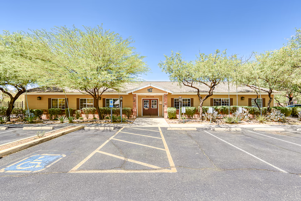 Front exterior of a single-story memory care building with a central entrance, shaded trees, and a parking lot with handicap spaces.