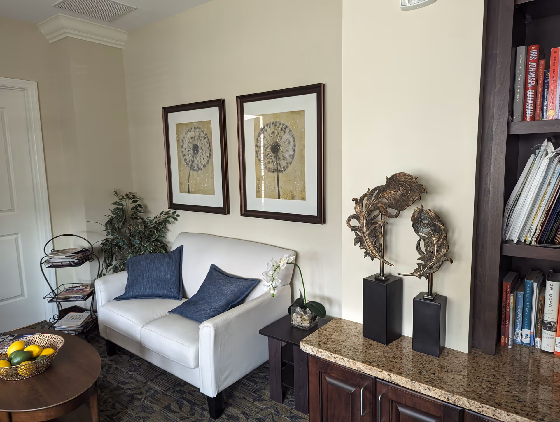 A cozy living room area with a white loveseat adorned with two blue cushions. Above the loveseat, there are two framed pictures of dandelions. To the right, there is a granite countertop with two decorative leaf sculptures on black stands. A dark wooden bookshelf filled with books and magazines is partially visible. On the left side, there is a small metal rack holding magazines and a green leafy plant next to the loveseat. A round wooden coffee table with a basket of lemons and limes is in the foreground.