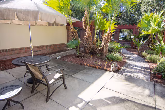 Outdoor patio area with a round table, two chairs, and a large umbrella. The patio is adjacent to a garden with palm trees and a brick pathway leading through the greenery.
