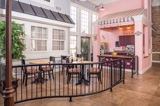 Indoor seating area with several round tables and chairs with floral cushions, separated by a black metal railing. To the right, there is a pink concession stand or snack bar with a striped awning, popcorn machine, and coffee equipment. Large windows and a door are visible in the background, along with a decorative white structure resembling a small house facade.