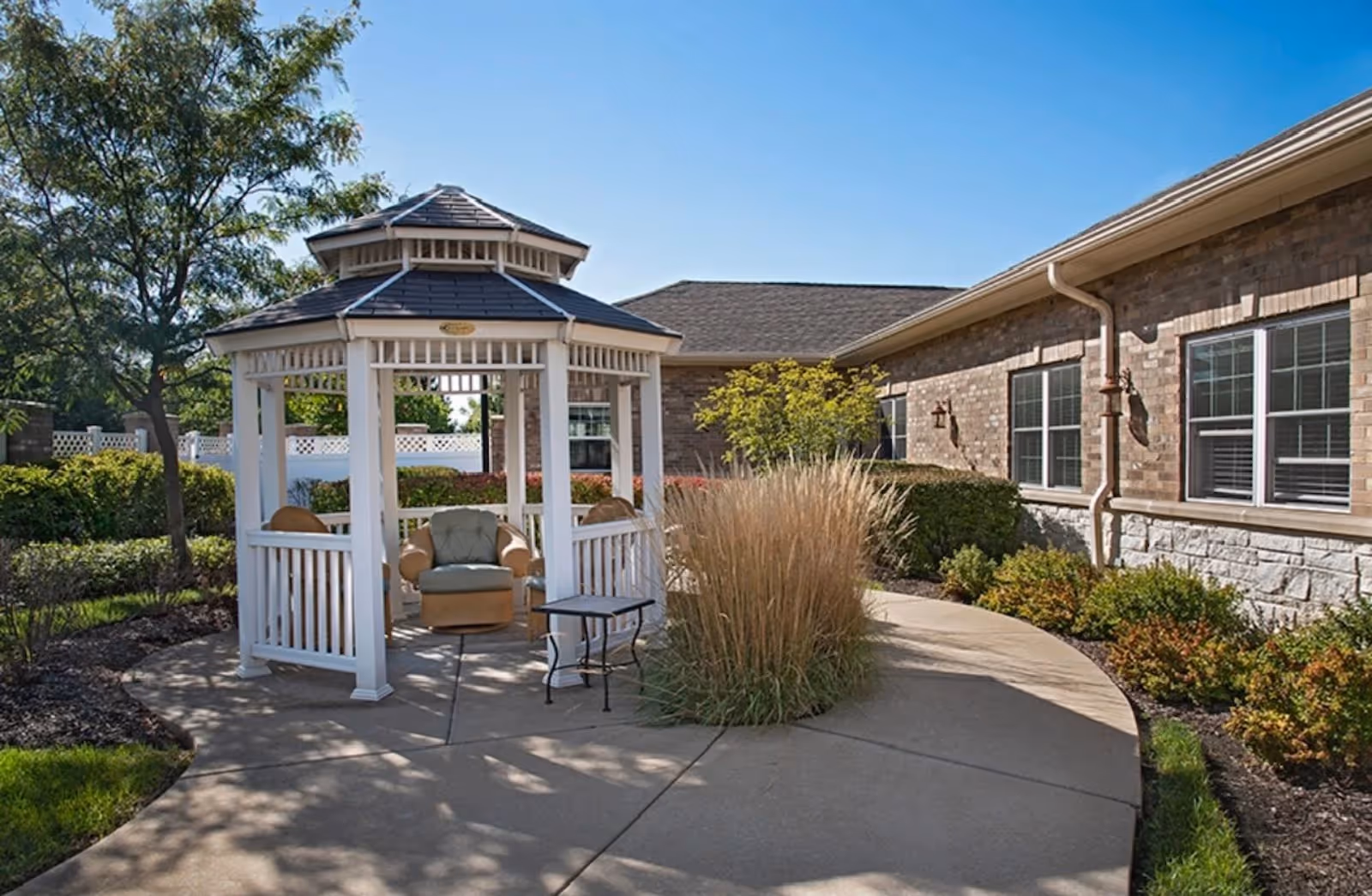 Outdoor garden area at a senior living facility featuring a white gazebo with cushioned chairs and a small table, surrounded by landscaped bushes, ornamental grasses, and a brick building with windows in the background under a clear blue sky.