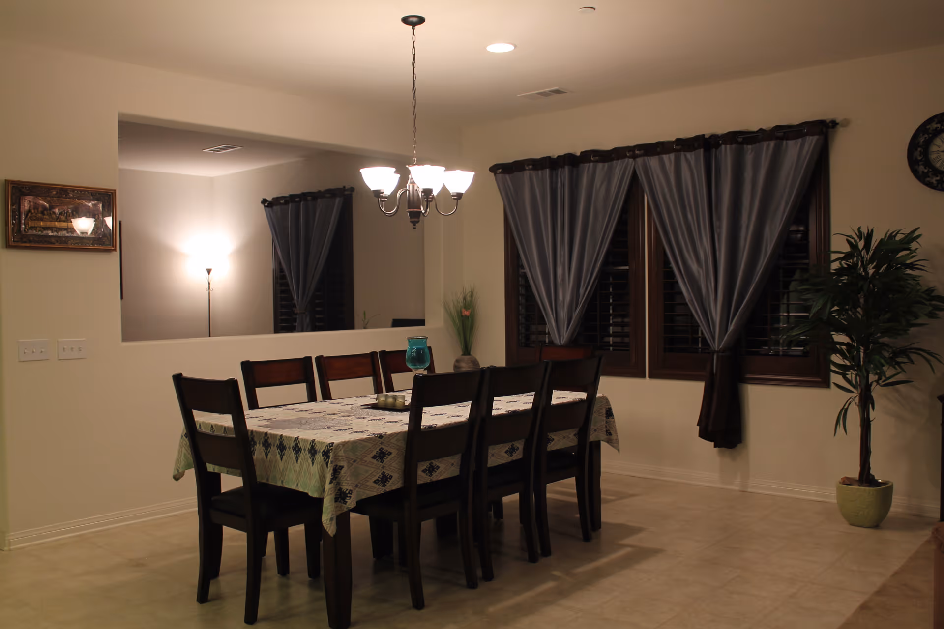 A dining room with a rectangular table covered with a patterned tablecloth and surrounded by eight wooden chairs. Above the table hangs a chandelier with five lights. The room has beige walls, tiled floor, and three windows with dark curtains. A potted plant is placed near the windows, and a wall clock is visible on the right side. There is a large interior window opening to another room with a floor lamp and framed artwork on the wall.