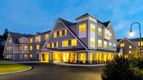Exterior view of a large, well-lit senior living facility building at dusk with multiple windows glowing warmly. The building has a modern design with gray siding, white trim, and a covered entrance. The driveway curves in front of the entrance, and there is some landscaping with bushes and grass around the building.