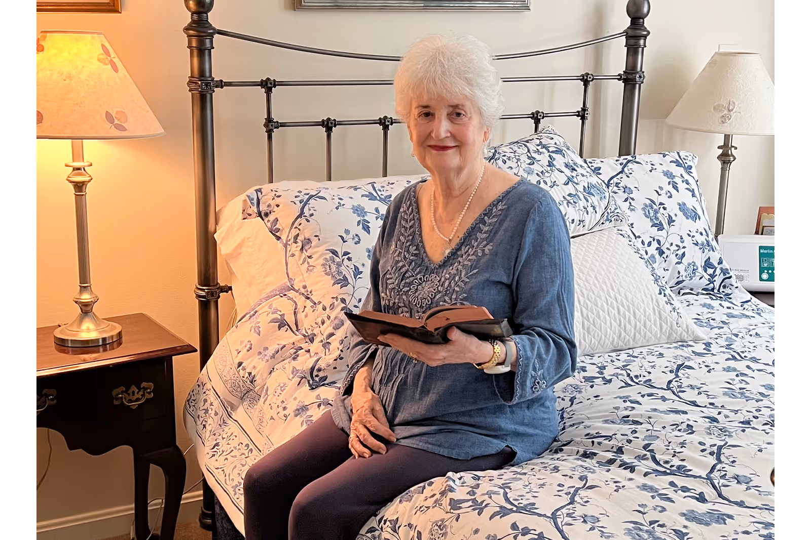 An elderly woman with white hair sitting on a bed with blue and white floral bedding, holding an open book. The room has a metal bed frame, two bedside tables with lamps, and a cozy, well-lit atmosphere.