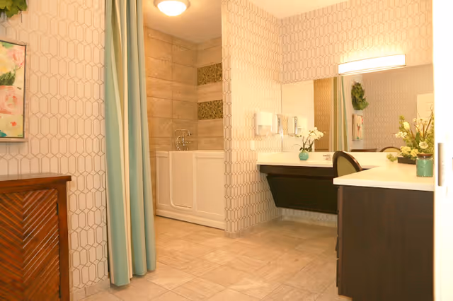 A spacious bathroom featuring a walk-in bathtub with tiled walls and decorative tile accents. There is a large vanity with a wide mirror, a chair, and floral decorations on the countertop. The walls have a geometric patterned wallpaper, and a green curtain partially covers the bathtub area.