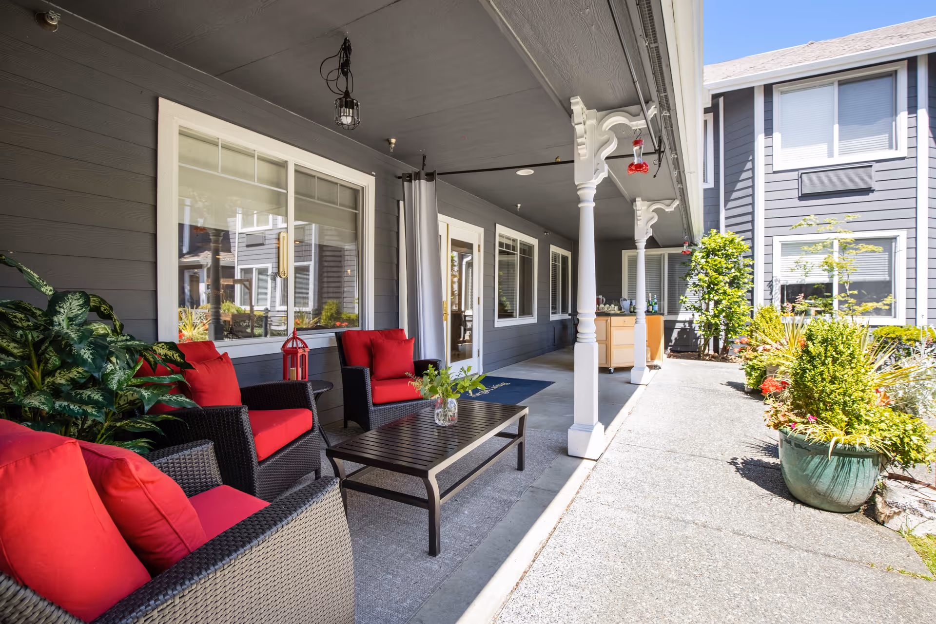 Outdoor covered patio area with dark wicker furniture featuring red cushions, a black coffee table with a small plant, and decorative plants along the side. The building exterior is gray with white trim, and there are white columns supporting the roof. The patio opens to a walkway with more greenery and potted plants under a clear blue sky.