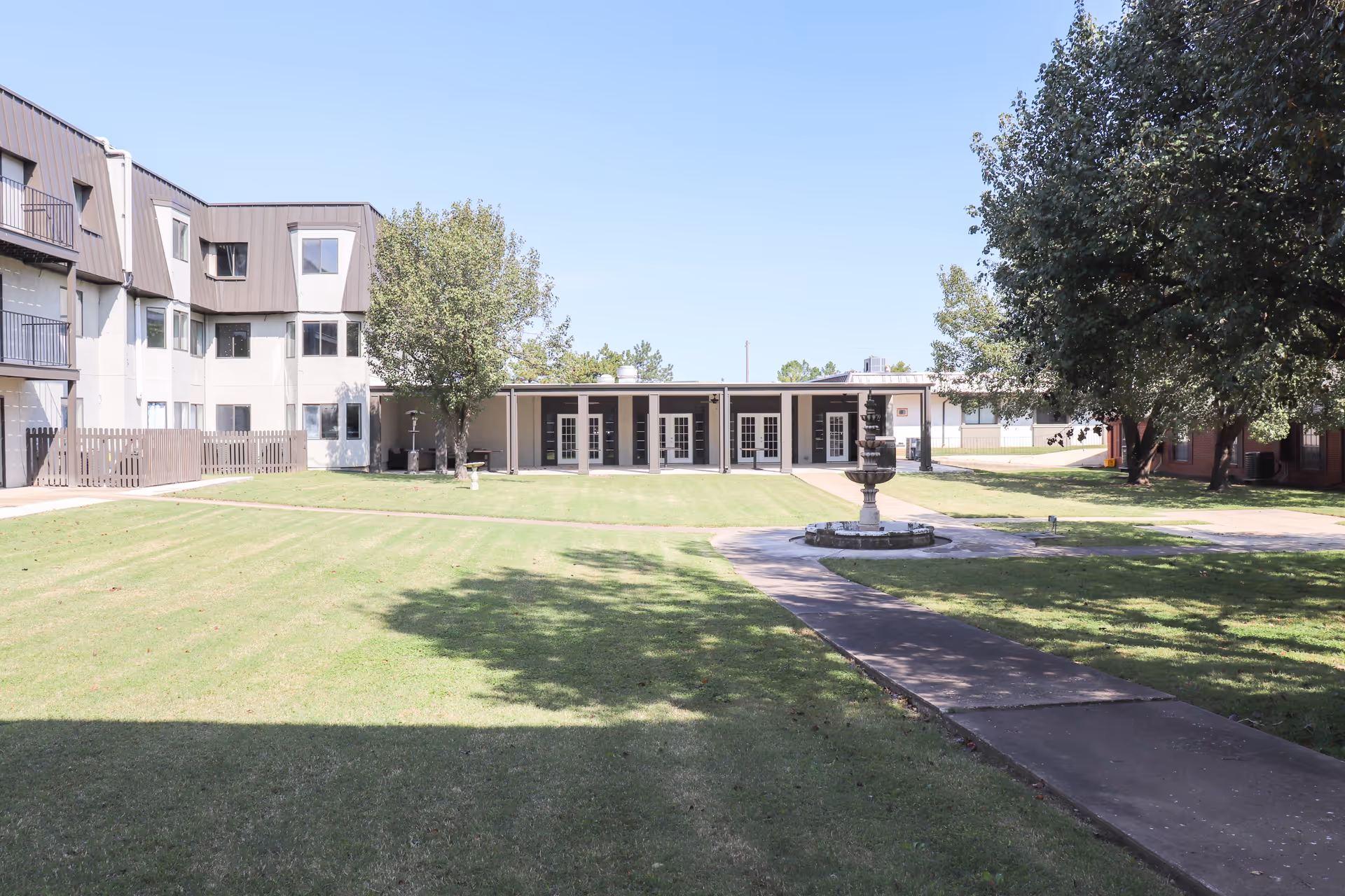 Outdoor view of Southern Hills Retirement Community showing a well-maintained grassy courtyard with a paved walkway leading to a central fountain. Surrounding the courtyard are multi-story residential buildings and trees under a clear blue sky.