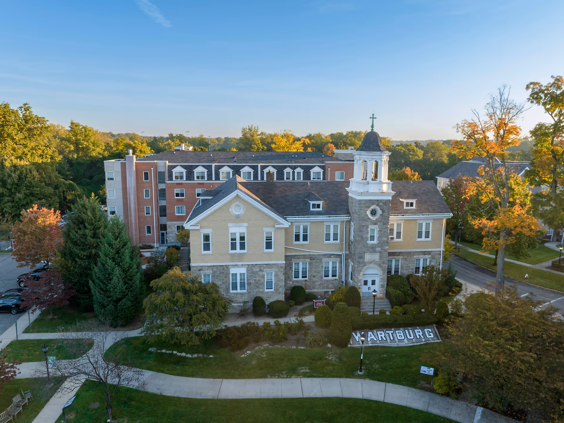 Aerial view of The Wartburg Home building surrounded by trees with autumn foliage. The building has a stone and beige facade with a tower featuring a cross on top. There is a well-maintained lawn and pathways in front of the building, with a sign that reads 'WARTBURG'.